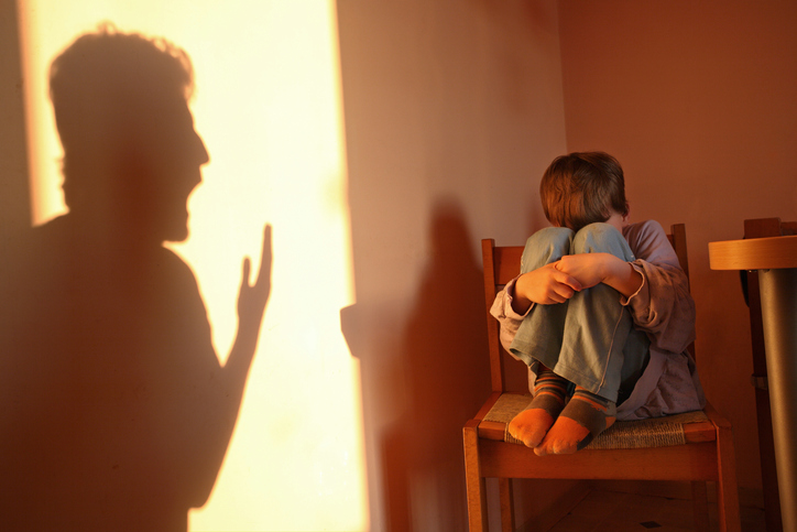 A boy cowers in a dark corner. A yelling adult casts a shadow on the wall.