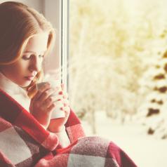 Person with long auburn hair sits in window, wrapped in red and white plaid blanket, inhaling steam from hot drink